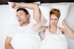 Couple in bed, man snoring loudly while woman covers her ears, illustrating the impact of sleep apnea on partners and the importance of treatment.