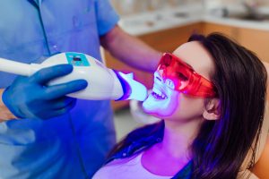 Patient undergoing in-office teeth whitening treatment with LED light activation, wearing protective eyewear, and receiving care from a dental professional.
