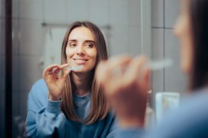 Woman smiling while applying teeth whitening treatment in front of a bathroom mirror, illustrating at-home whitening options.